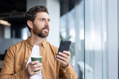Elinde telefon ve kahve fincanı olan sakallı bir adam ofis ortamında pencereden dışarı bakıyor..