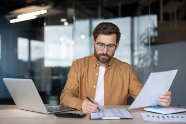 Çalışma masasında bir işadamı dizüstü bilgisayarda çalışırken belgeleri inceliyor. Etrafı çizelgeler, kağıtlar ve hesap makinesiyle çevrili..