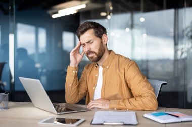 Stresli bir işadamı masasında oturuyor, eli alnında. Bir dizüstü bilgisayar, belgeler ve tablet masada..