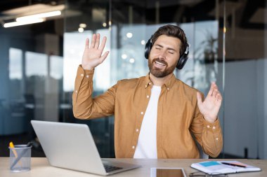Bir adam masasında otururken kulaklıkla müzik dinlemekten zevk alır, ellerini kaldırarak bir neşe ve minnettarlık hareketi yapar..