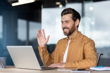 Bir video görüşmesi sırasında gülümseyen bir iş adamı, modern ofis ortamında oturuyor, bir masanın üstünde dizüstü bilgisayar kullanıyor..