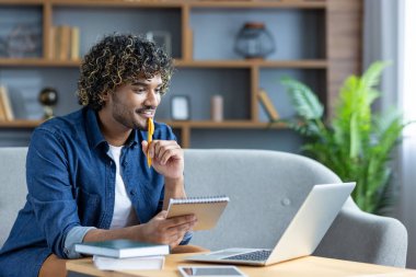 Genç bir adam evde çalışıyor, dizüstü bilgisayarına bakarken not alıyor. Düşünceli görünüyor..