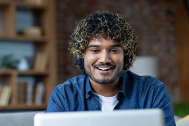 Kıvırcık saçlı, kulaklık takan, evden çalışan gülümseyen bir adam. O sıcak bir ifadeyle kameraya bakıyor..