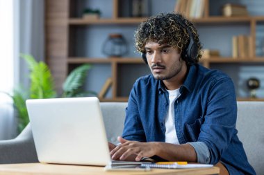 Kulaklıklı genç bir adam evden bilgisayarda çalışıyor, sanal bir toplantıya katılıyor ya da uzaktan çalışıyor..