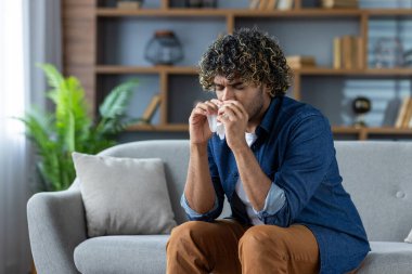 Kanepede oturan kıvırcık saçlı bir adam, burnunu siliyor, muhtemelen mevsimsel alerji ya da soğuk algınlığı geçiriyor..