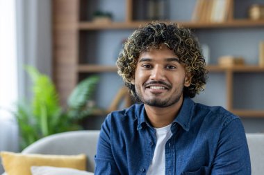 Kıvırcık saçlı ve sakallı gülümseyen genç bir adam fotoğraf için içeride poz veriyor, doğrudan kameraya bakıyor..