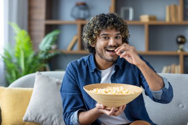 Gülümseyen bir adam evdeki kanepede dinlenirken patlamış mısırın tadını çıkarır. Mutlu ve abur cubur yiyor..