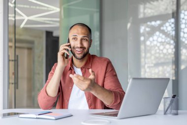 Gülümseyen bir işadamı, telefonuyla parlak bir ofis ortamında dizüstü bilgisayarla konuşurken el kol hareketi yapar..