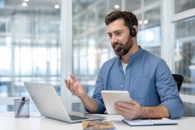 Kulaklık takmış bir adam elinde tablet, elinde dizüstü bilgisayar, gözlük ve defterlerle bir masada otururken el kol hareketi yapar..
