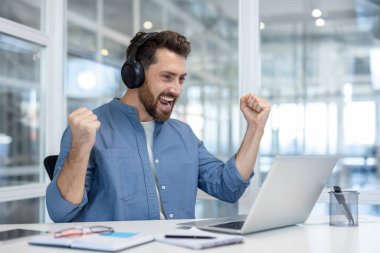 Bir adam zaferi kutlar, kulaklık takar ve ofis ortamında dizüstü bilgisayarına bakar. Heyecandan yumruklarını sıkıyor..
