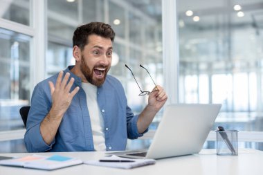 Ofis ortamında dizüstü bilgisayarına bakan sakallı, şaşırmış bir adam, gözlüğünü hayret içinde tutuyor..