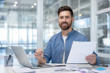 Mavi gömlekli gülümseyen bir işadamı, modern bir ofiste, masasında dizüstü bilgisayar ve gözlüklerle belgeleri gözden geçirir..