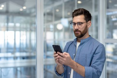 Gözlüklü bir adam ofis ortamında telefonuna bakıyor, muhtemelen mesajlaşıyor ya da bakınıyor..