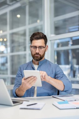 Gözlüklü bir adam modern ofis ortamında masasında tablet kullanır..