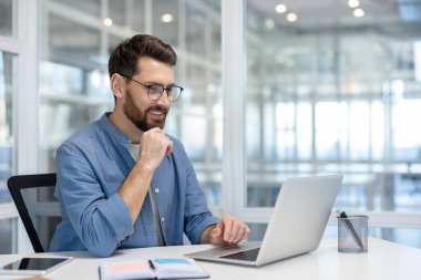 Profesyonel bir adam dizüstü bilgisayarda çalışıyor, gülümsüyor, parlak, modern ofis ortamında..