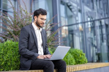 Gözlüklü bir işadamı, modern bir bina ve yeşillik yakınlarındaki açık havada bilgisayarında çalışıyor..