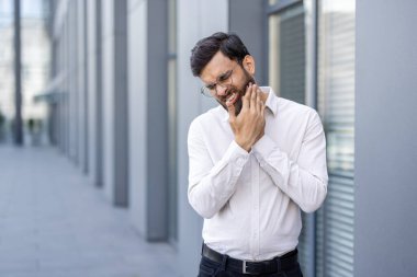 Beyaz gömlekli bir adam, modern bir binanın yanında çenesini açık tutarken acı hisseder..
