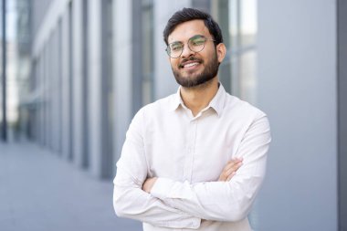 Gülümseyen, kendine güvenen gözlüklü bir Hintli adam dışarıda duruyor, kollarını kavuşturuyor, bulanık bir modern mimari arka plana karşı..