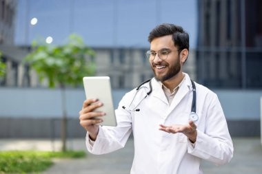 Beyaz önlüklü gülümseyen bir doktor, kliniğin dışında bir tablet kullanıyor..