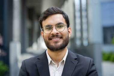 Sakallı ve gözlüklü, güler yüzlü bir işadamı, bir binanın önünde, takım elbise giymiş, kendinden emin bir şekilde duruyor..