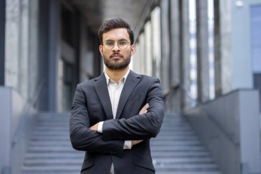 Takım elbiseli bir Hintli işadamı modern bir binanın önünde kollarını kavuşturmuş duruyor..