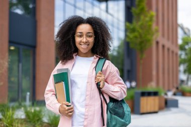 Bir üniversite binasının önünde elinde kitap ve sırt çantası olan mutlu bir Afrikalı Amerikalı öğrenci gülümsüyor..