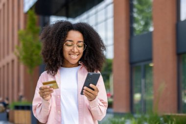 Gözlüklü genç bir kadın telefonunu kullanırken ve elinde kredi kartı tutarken gülümsüyor, muhtemelen internetten alışveriş yapıyor..