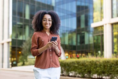 Kıvırcık saçlı gülümseyen bir kadın gözlük takar ve güneşli bir günde modern bir binanın önünde akıllı telefon kullanır..