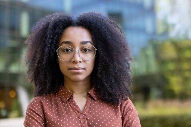 Kıvırcık saçlı, gözlüklü ve açık havada yakalı bir gömlek giyen genç bir kadının odaklanmış portresi..