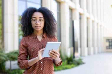 Gözlüklü genç bir kadın bir ofis binasının önünde tablet tutuyor..