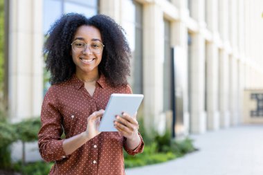 Gözlüklü genç bir kadın gülümserken tablet tutuyor. Arka planda bulanık bir bina var..