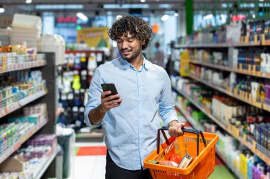 Marketten alışveriş yapan bir adam, elinde alışveriş sepeti tutarken telefonunu kullanıyor. Gülümsüyor..