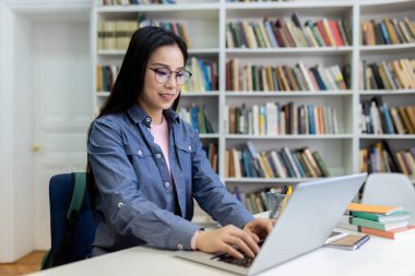 Kütüphanede dizüstü bilgisayar kullanan, arka planda kitaplığı olan bir kadın. Ders çalışıyor..