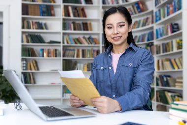 Gülümseyen Asyalı bir kadın kütüphane ortamında belgeler tutuyor. Kitaplıklarla dolu kitaplıklar bir arkaplan oluşturur.