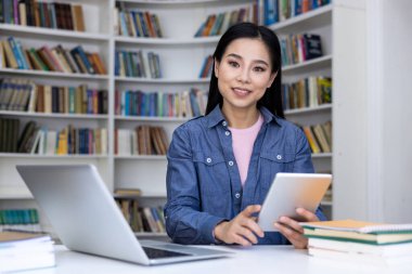Asyalı bir kadın elinde tablet tutarken gülümsüyor. Kütüphanede çalışıyor. Yanında dizüstü bilgisayarı ve kitapları var..
