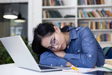 Kütüphanedeki bir masada uyuyan yorgun bir öğrenci. Yanında dizüstü bilgisayar ve kitaplarla..
