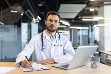 Beyaz önlüklü yakışıklı bir doktor masasında oturuyor, önünde bir dizüstü bilgisayarla bir panoya yazı yazıyor. Kameraya özgüvenle bakıyor..
