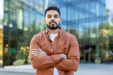 Sakallı, kendine güvenen bir adam modern bir ofis binasının önünde kollarını kavuşturarak poz veriyor. Profesyonellik ve ulaşılabilirlik yayıyor..