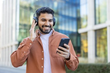 Kulaklık takan ve şehir merkezinde akıllı telefonuna bakan gülümseyen bir adam..