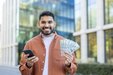 Elinde para ve telefon olan gülümseyen bir adam modern bir binanın önünde dikiliyor..