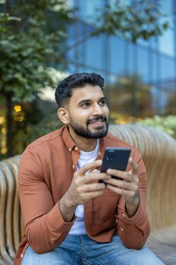 Gülümseyen bir adam parktaki ahşap bir bankta otururken telefonuna bakar..