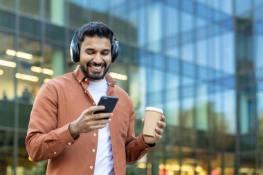 Gülümseyen bir adam modern bir binanın önünde telefonunu kullanırken müzik ve kahveden hoşlanır..