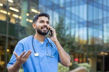 Bir sağlık uzmanı telefonla iletişim kurar, modern bir binanın yakınında el kol hareketi yapar, muhtemelen hasta bakımını ya da tıbbi prosedürleri tartışır..