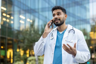 Laboratuvar önlüğü giyen ve steteskop ile konuşan bir doktor modern bir binanın yakınında el kol hareketi yapıyor..