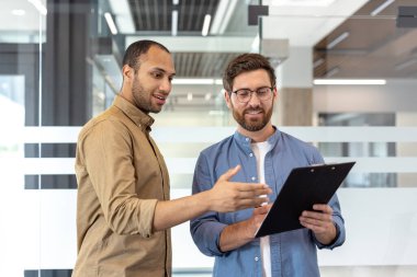 İki farklı iş adamı, belgeyi modern bir ofis ortamında, muhtemelen ortak bir toplantı veya sunum sırasında tartışıyor..