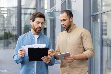 İki iş ortağı modern bir binanın önünde belgeleri gözden geçiriyorlar, bir proje üzerinde çalışıyorlar..