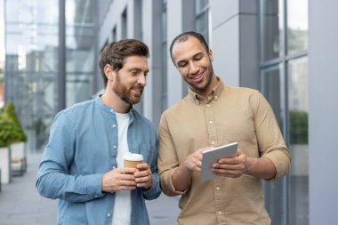 İki işadamı, biri kahve fincanı tutuyor, tablete bak. Modern bir binanın dışında bir şey tartışıyorlarmış gibi görünüyor..