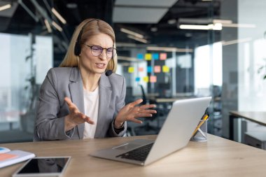 Gergin ve sinirli bir kadın ofiste oturmuş video görüşmesi için kulaklık ve dizüstü bilgisayar kullanıyor. İş kadını takım elbiseli online iş konferansında.