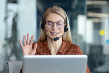 Kulaklık takan, gözlüklü bir kadın modern ofis ortamında dizüstü bilgisayara bakarken el sallıyor..