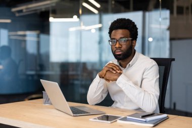 Profesyonel ortamda gözlüklü bir adam, masada dizüstü bilgisayarla çalışıyor..